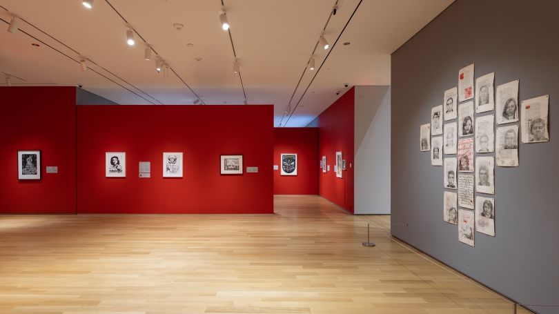 A museum gallery with mostly red walls, but the wall to the right is painted a dark grey. On the gray wall, in a diamond pattern, hang unframed works on paper. These works are printed with portraits over text, which appear to be official records.
