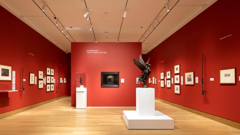 A museum gallery with red walls and white wall text. There are framed prints hanging on the wall. In the middle of the room is a sculpture of a winged figure carrying away another figure.