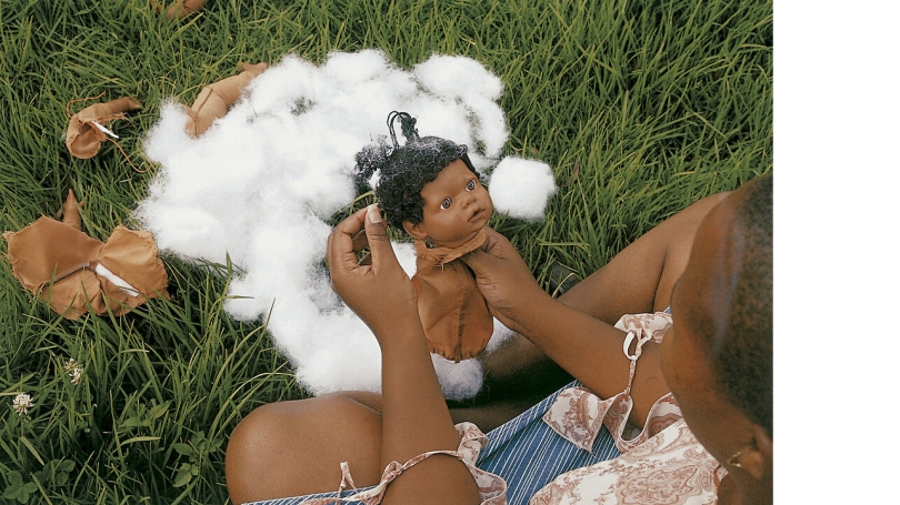 A photograph of a Black woman (from above). She is sitting in a field and the focus of the photo is on her hands which are holding and creating a Black baby doll.