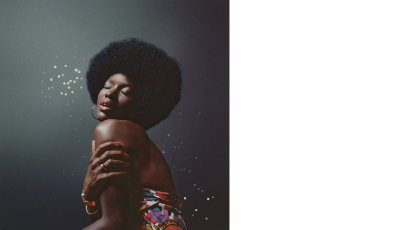 A beautiful dark skinned woman with an afro stands in profile to the camera, but her face is turned and in full view. Her eyes are closed and her face is titled slightly up. She has a calm or serene expression. Blurred lights behind her sparkle.
