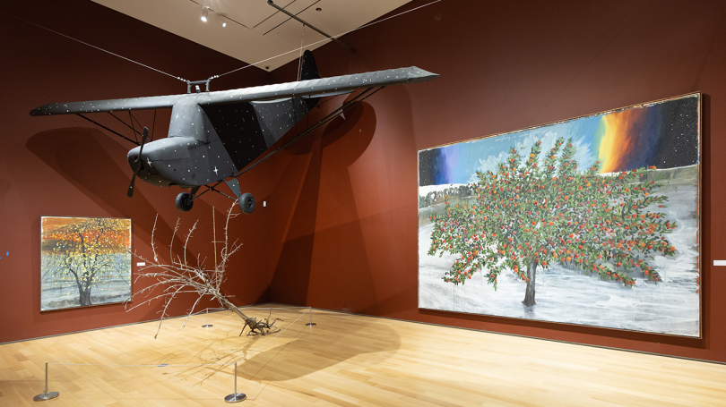  View of "The Grief of Almost," installed in Northeast Gallery. The walls are painted red, and there is a large plane hung from the ceiling surrounded by art on the walls. 