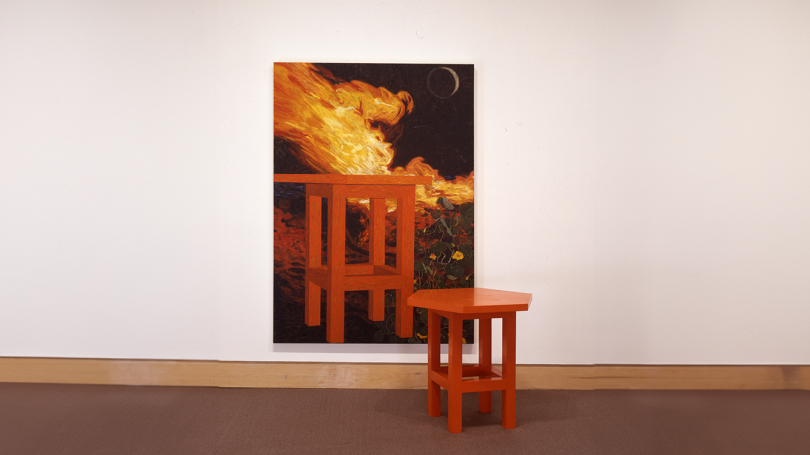 A red hexagonal table sits infront of a painting of the same table. In the painting, the table is on fire and is set in front of a nigh time sky. 