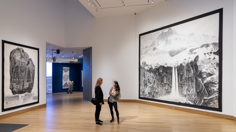 Two women stand in a museum gallery surrounded by gigantic works on paper of mountain landscapes.