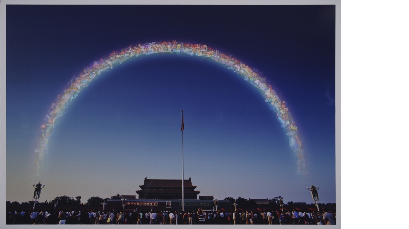 A photograph of a Chinese town or city. People are gathered in the foreground and traditional Chinese architecture is visible in the background. The people gathered are looking up at a rainbow that is actually made of stickers.