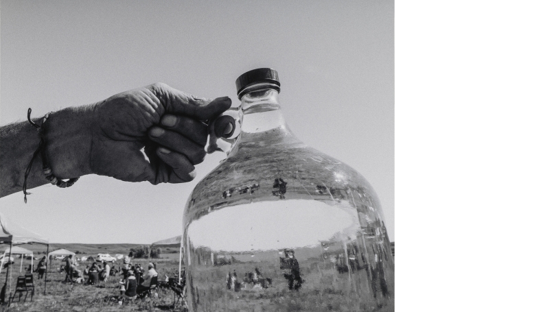 A black and white photograph of a hand holding out a large glass container. Half of the photo is viewed through the glass bottle and the other half is not. The location appears to be a cattle farm.
