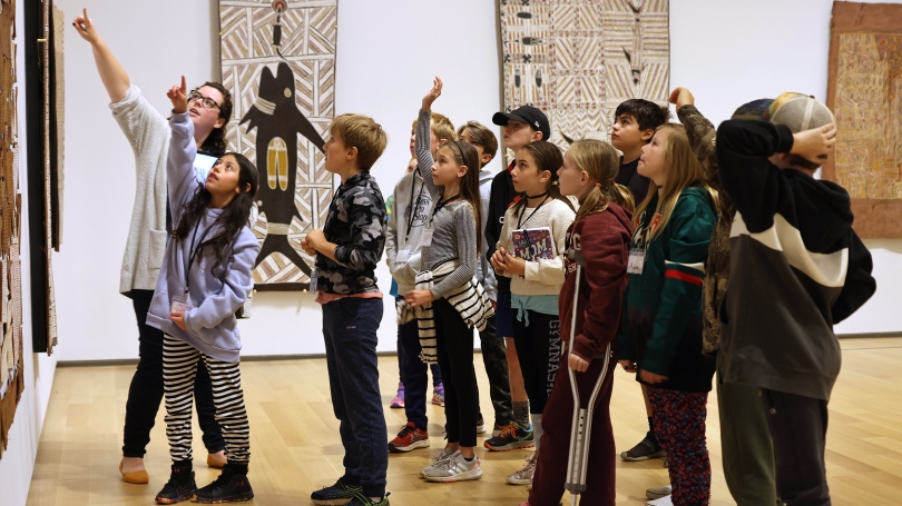 A group of elementary school children stand grouped in a half circle and are pointing up at something in a work of art. They are looking at Australian Aboriginal Bark Paintings.