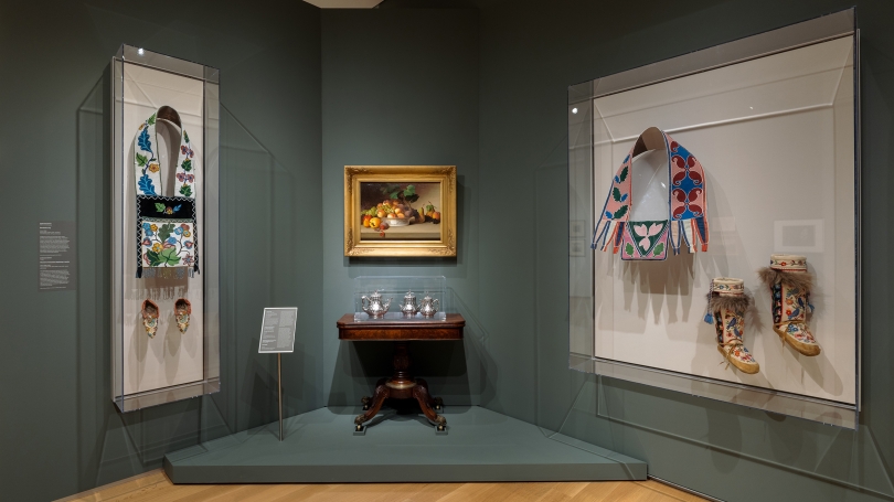 A museum gallery niche wall with wall cases containing traditional beaded Native American clothing and bags, a decorative arts table, a silver set, and a still life painting of a bowl of fruit.