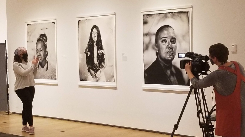 A college student discusses an exhibition virtually with her fellow classmates. She is standing in a museum gallery, surrounded by large black and white portraits, and being filmed by a museum staff member.