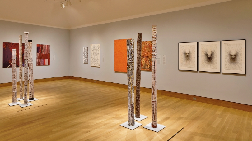 A museum installation of Indigenous Australian art. In the center of the gallery are two groupings of painted poles, and they are surrounded by various types of Indigenous Australian art (prints, paintings, etc).