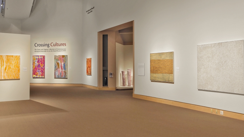 A large museum gallery with carpet, white walls, and a high ceiling, displaying paintings on canvases by Indigenous Australian artists.