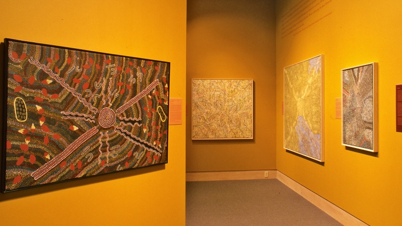A museum gallery with golden yellow walls and works by Indigenous Australian painters hang on the walls. 