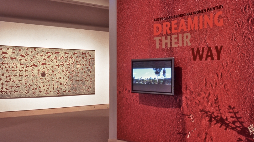 A museum gallery displaying works by Indigenous Australian artists. To the right in the photograph, is a curved wall with vinyl that displays the exhibition title and looks like red sand, which also holds a TV. To the left is a large horizontal painting.