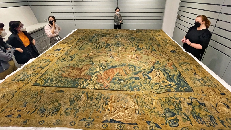 Four adults stand around a large light gray table that has a 16th century tapestry laid upon it. The tapestry is gold, blue green, and depicts the scene of a great hunt.