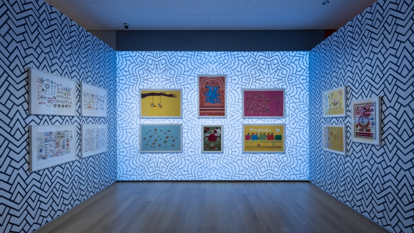 View of a gallery with a series of 13 prints. The lighting is slightly blue and the walls have a black-and-white zigzag pattern projected on them. 