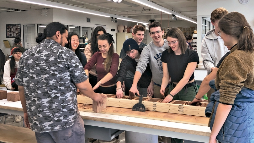 A photograph of a group of college students standing around a long table making clay bricks from a mold.