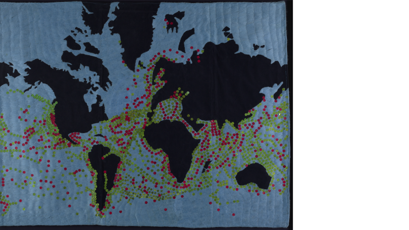 A woven map of the world. The continents are black, and the water is blue. Red and green sequins populate prominent shipping channels.  