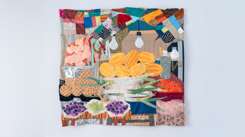 A colorful textile artwork depicting a vibrant market scene with various fruits and vegetables. The artwork includes quilted representations of melons, pineapples, and grapes, along with other produce items. The background features patchwork patterns and 