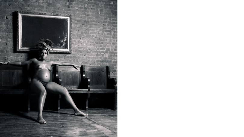 A topless and pregnant black woman sits on a bench and gazes at the camera. The photo is in black and white.