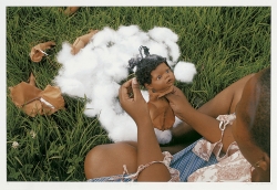 A photograph of a Black person is sitting on the ground in green grass. They are photographed from above and behind, the photographer is looking down into their lap where there is a Black baby doll being sewn together.