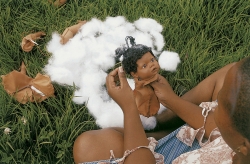 A photograph of a Black woman (from above). She is sitting in a field and the focus of the photo is on her hands which are holding and creating a Black baby doll.