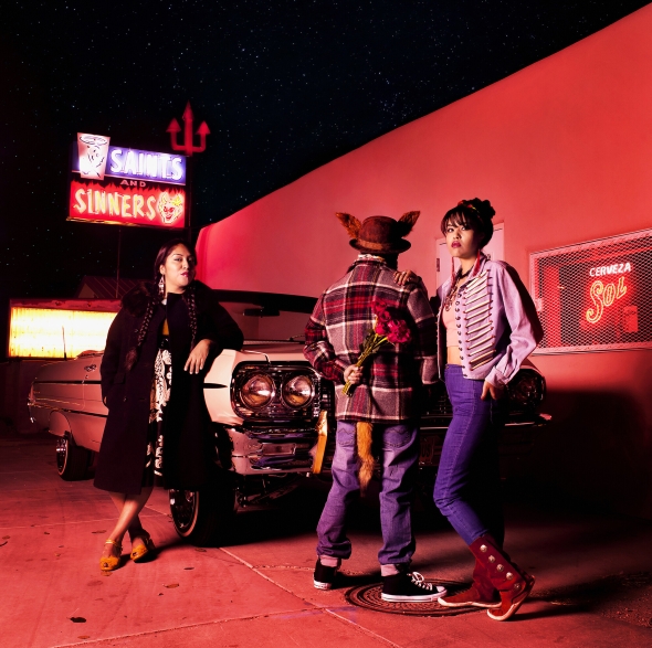 Three figures stand confidently near a vehicle. One is facing away from the cara and holding flowers behind their back.